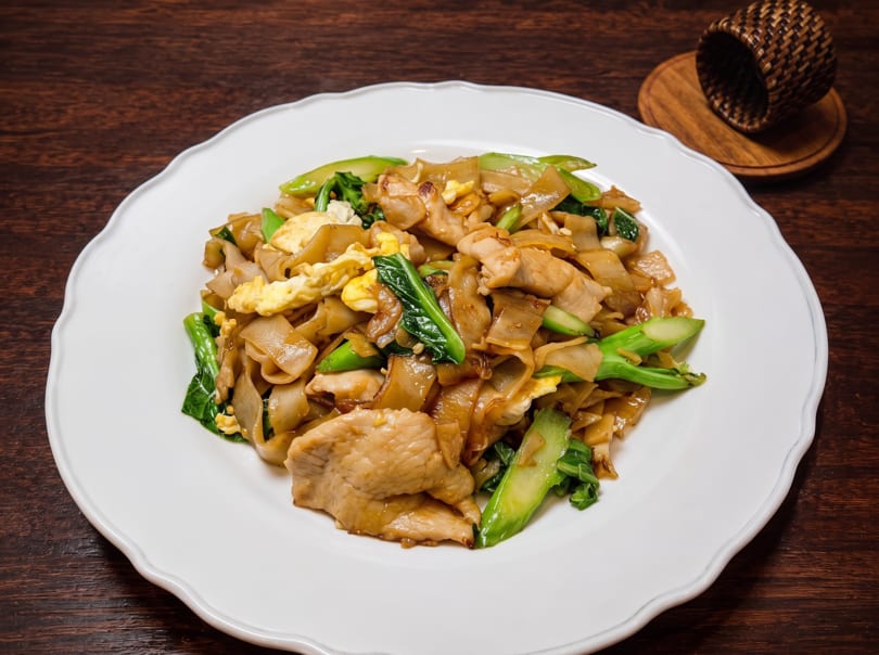 Pad See Ew with wide noodles, chicken, scrambled egg, and Chinese broccoli on a white plate.
