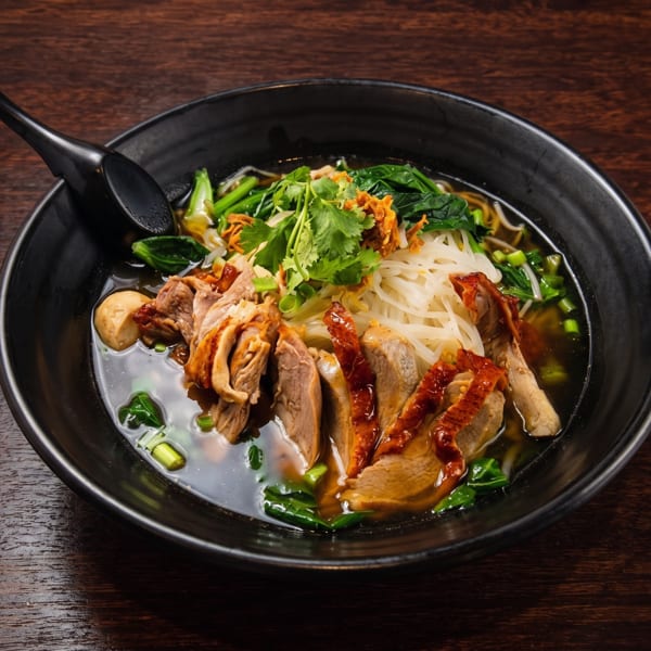 A black bowl of duck noodle soup with cilantro, greens, and a spoon on a wooden table.