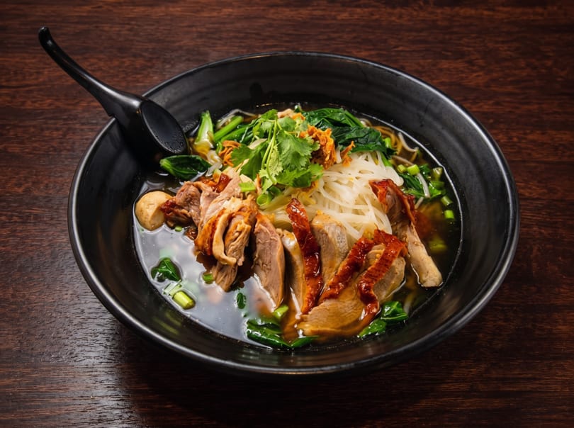 A black bowl of duck noodle soup with cilantro, greens, and a spoon on a wooden table.