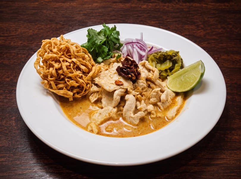 Khao Soi with chicken, crispy noodles, red onion, cilantro, pickled greens, and lime on a white plate.
