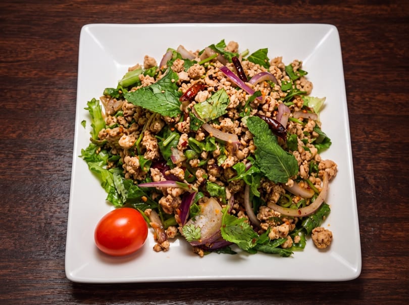 A white square plate of Thai Larb with minced meat, herbs, red onion, and a cherry tomato.