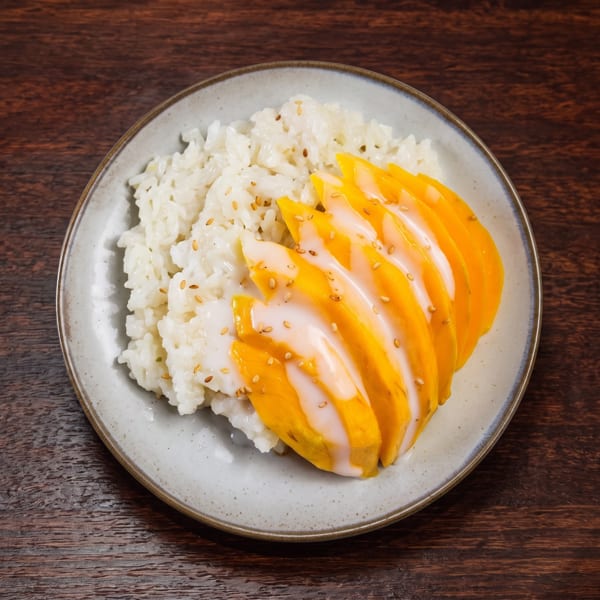 Mango sticky rice dessert with sesame seeds on a light gray plate over a dark wooden table.