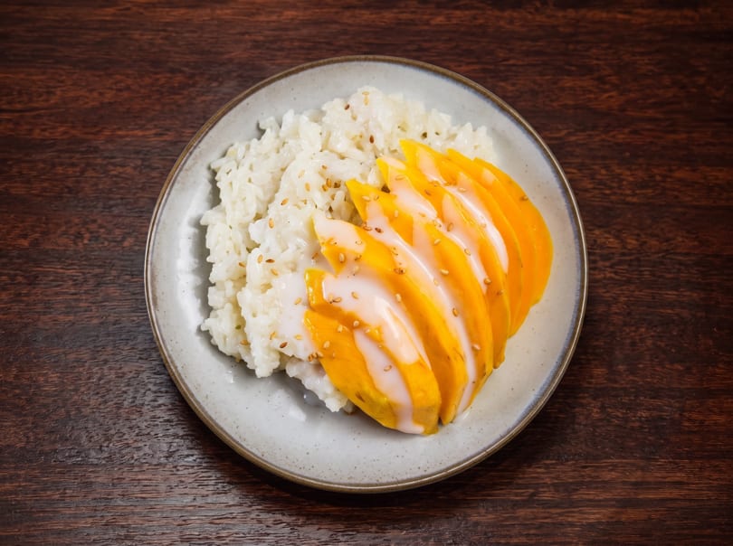 Mango sticky rice dessert with sesame seeds on a light gray plate over a dark wooden table.